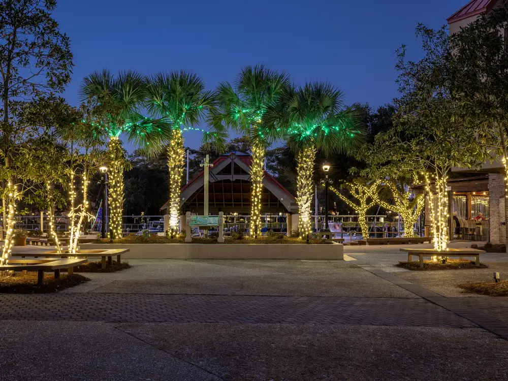 A commercial plaza at dusk with palm trunks wrapped in warm white lights and green lighting through the fronds