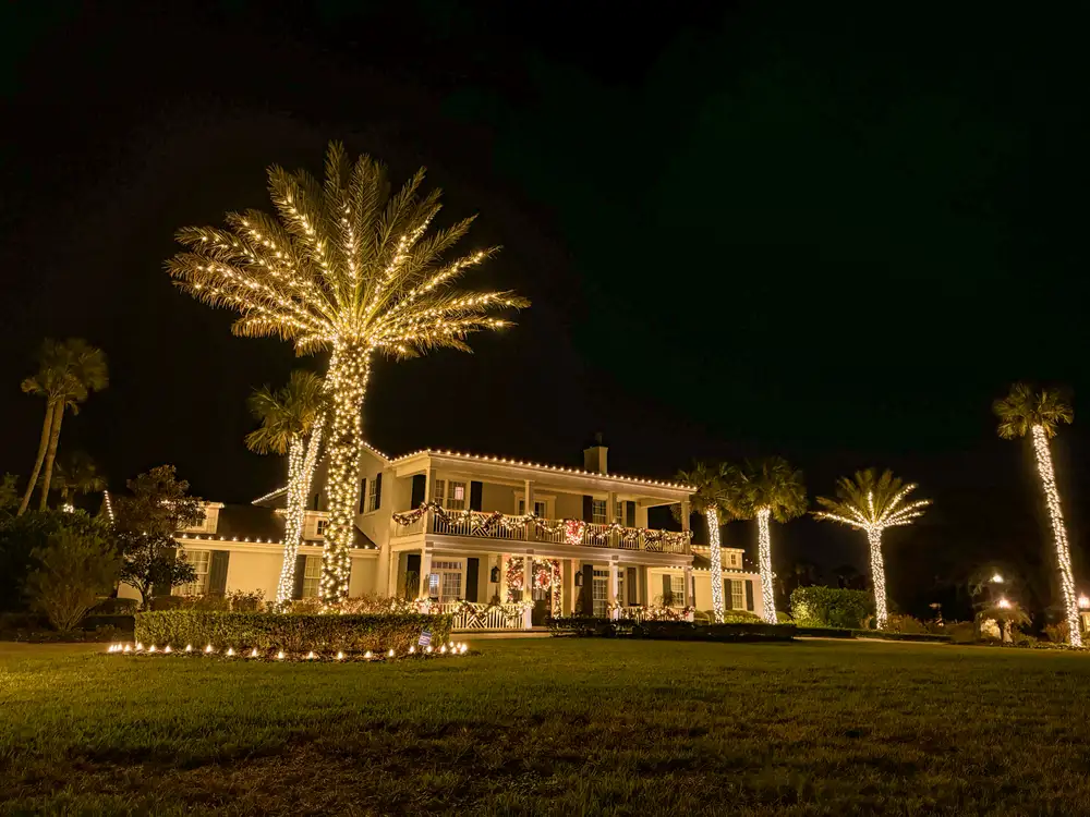 A Florida estate at night with roofline lighting, balcony garland and tall palms wrapped in warm white lights