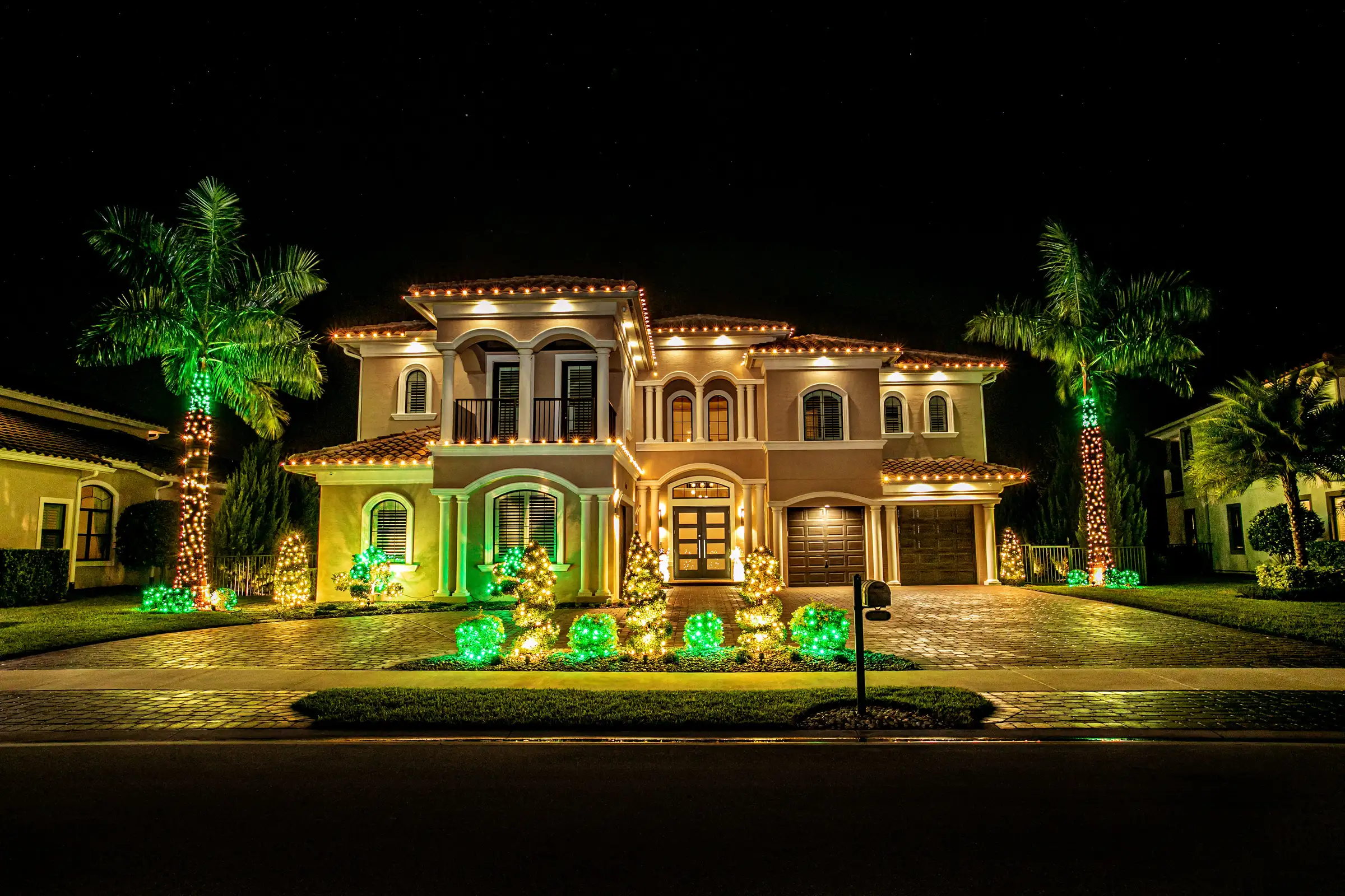 A Mediterranean-style Florida home at night with warm white lights tracing its rooflines, palms wrapped in lights, and green-lit shrubs across the front garden